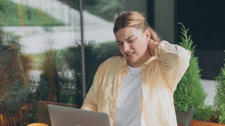 Femme d'une trentaine d'années souffrant de douleurs au cou, stressée, assise devant un ordinateur portable à l'extérieur